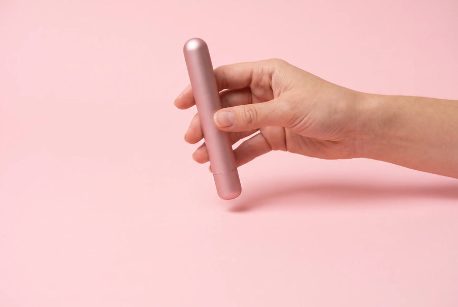 Een hand houdt een slanke roségouden massager tegen een zachte roze achtergrond